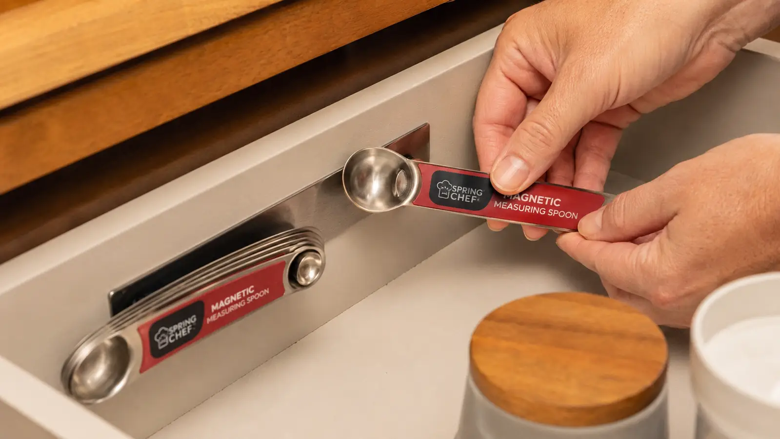 Hand attaching a stainless steel magnetic measuring spoon to a metal strip inside a kitchen drawer
