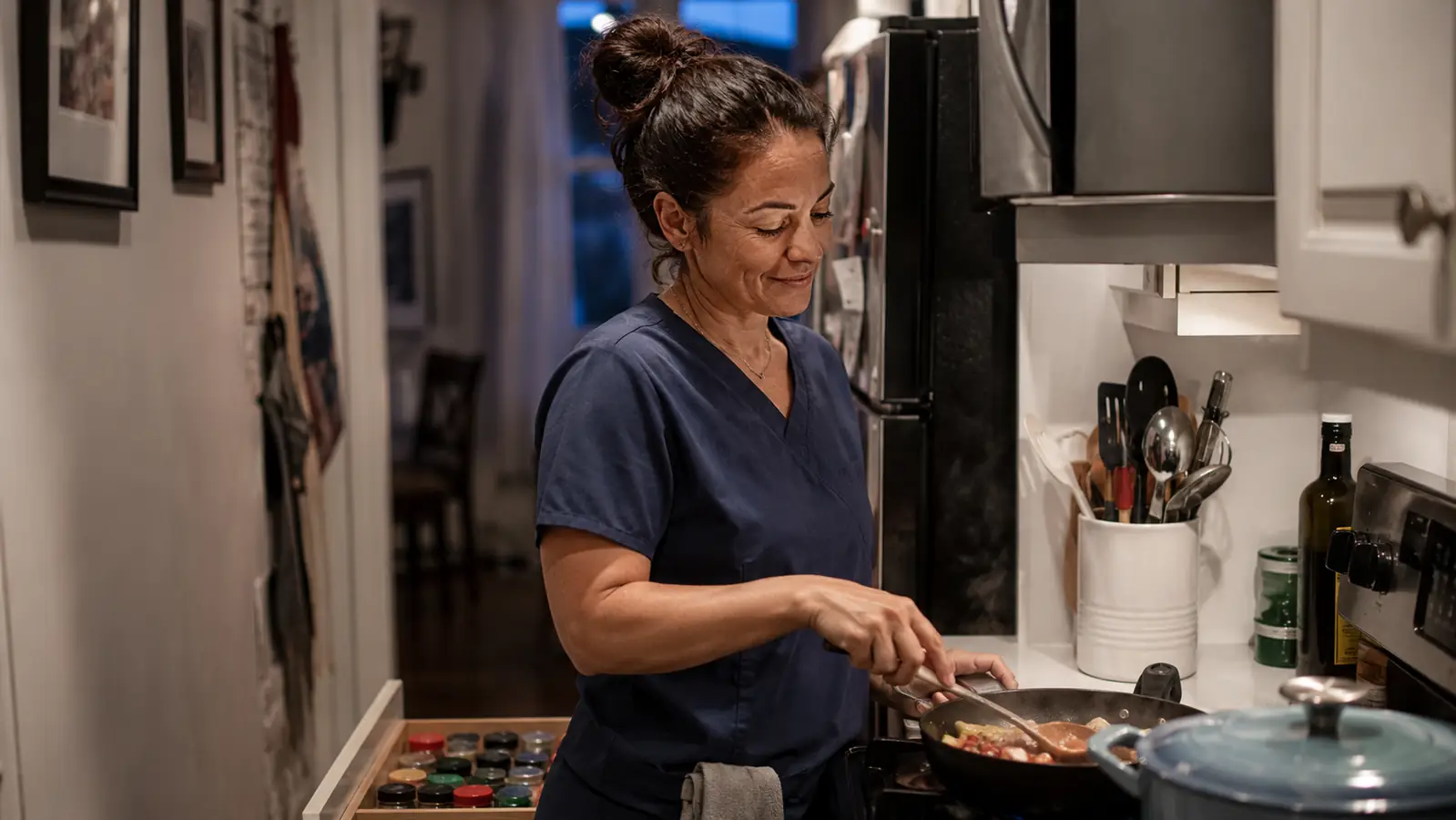 Woman in scrubs cooking dinner in a small kitchen after a hospital shift